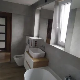 Nowoczesna łazienka z szarą glazurą, drewnianą podłogą i białymi sanitariatami, odbicie lustrzane ukazuje fragment dekoracji ściennej.