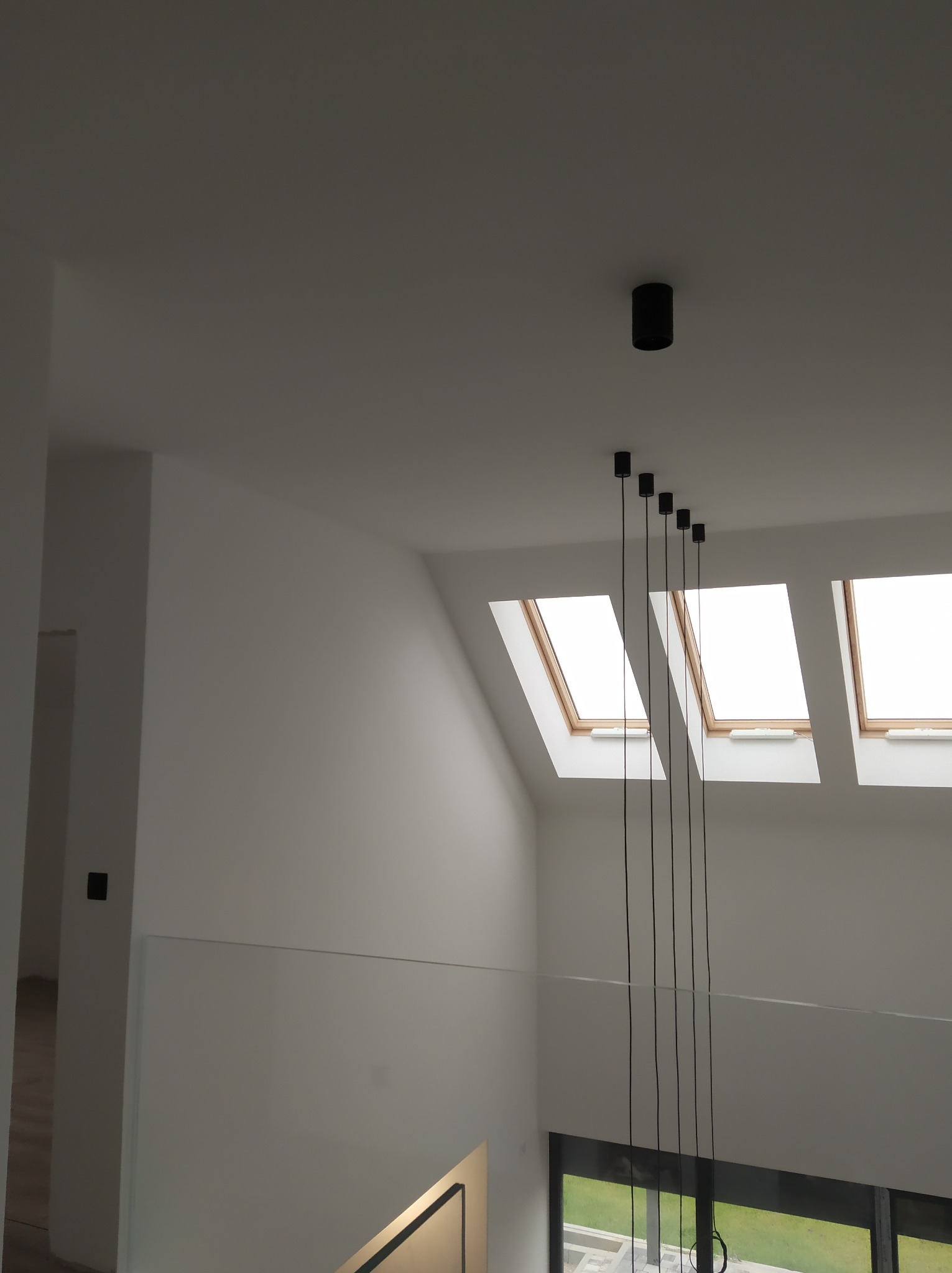 Nowoczesne wnętrze z białymi ścianami, szklaną balustradą i trzema oknami dachowymi; wiszące czarne lampy dodają minimalistycznego charakteru.