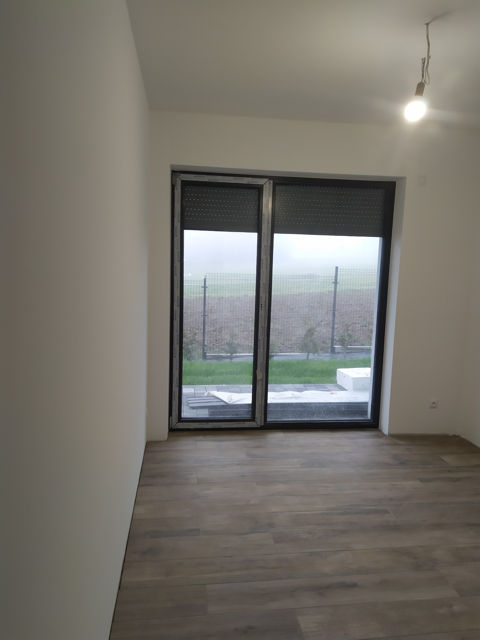 Pomalowane na biało wnętrze z drewnopodobną podłogą, widok na zewnątrz przez okno z roletami w pochmurny dzień.