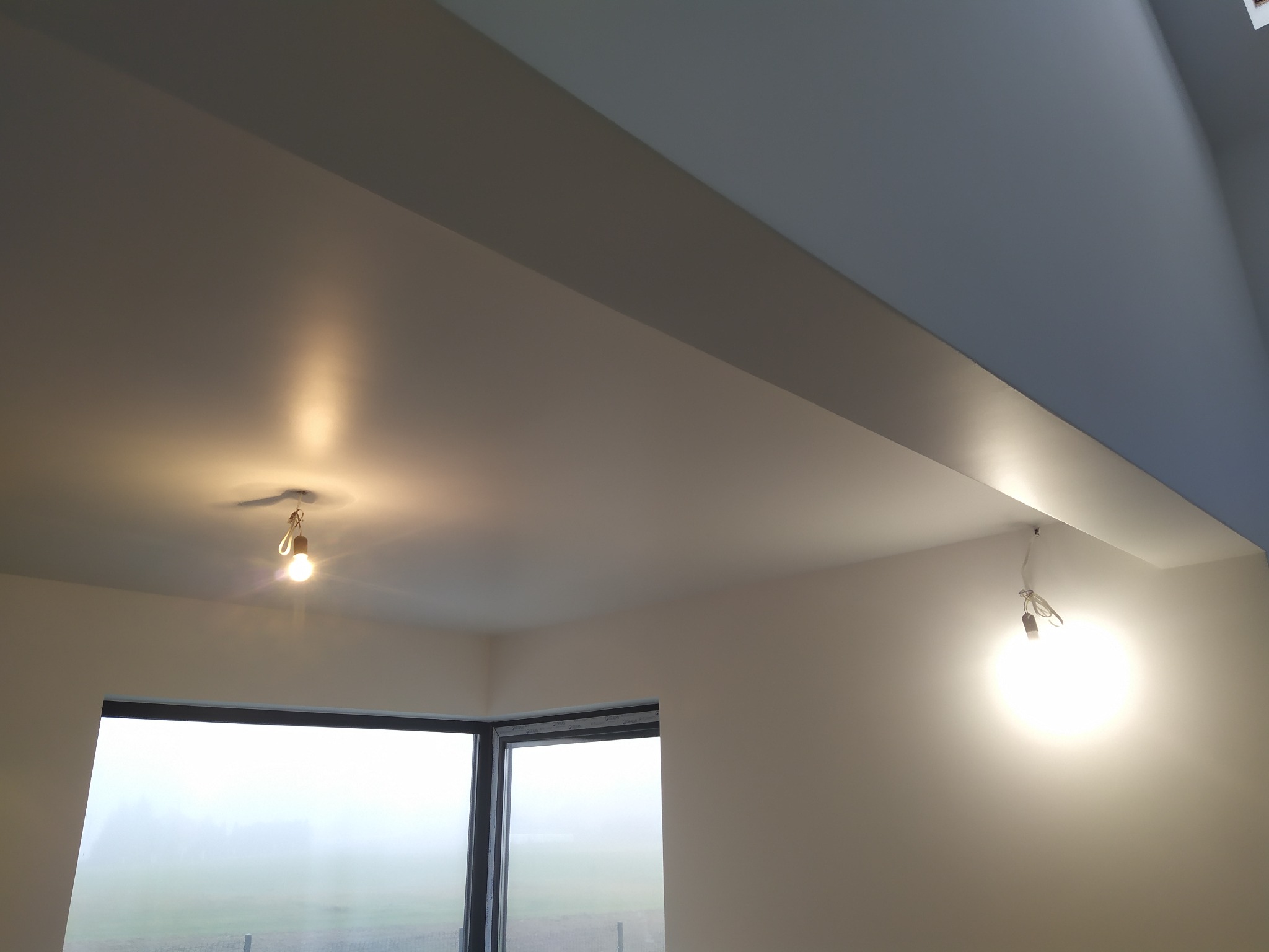 Pomieszczenie po remoncie, świeżo pomalowane ściany i sufit w jasnych odcieniach, duże okno z widokiem na zewnątrz w porannej mgle, dwie wiszące żarówki bez kloszy.