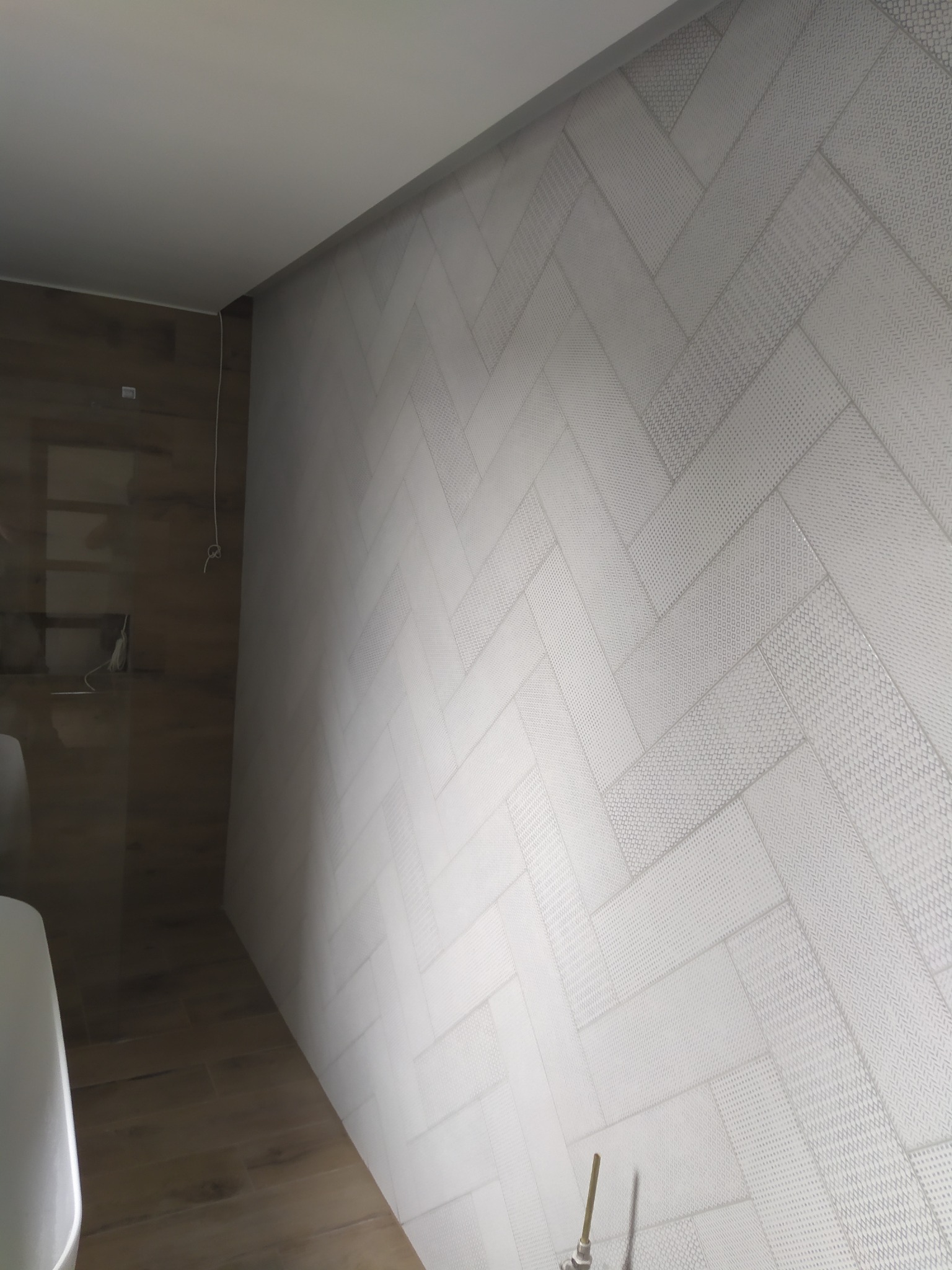 Ściana w łazience wyłożona białymi płytkami ułożonymi we wzór jodełki, z delikatnym wzorem na każdej płytce, oraz fragment brązowej ściany z widocznymi przewodami.