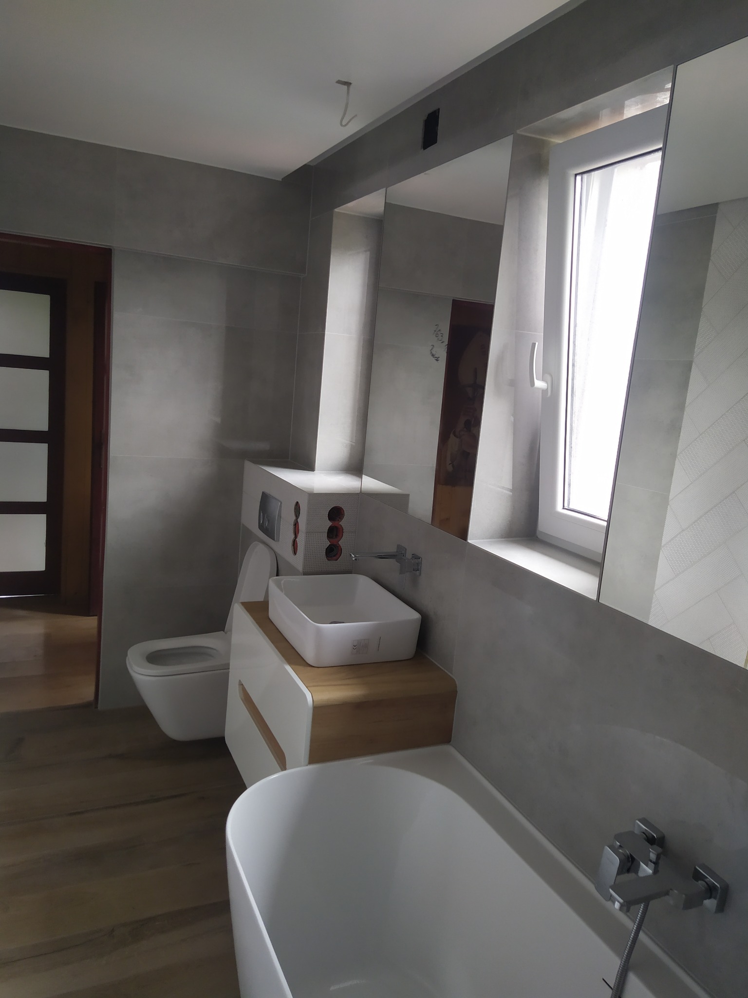 Nowoczesna łazienka z szarą glazurą, drewnianą podłogą i białymi sanitariatami, odbicie lustrzane ukazuje fragment dekoracji ściennej.