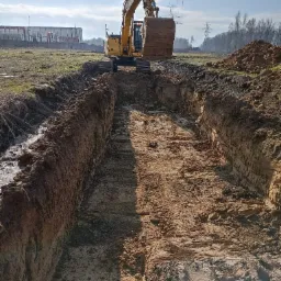 Żółta koparka na tle błękitnego nieba, wykonująca wykop liniowy w ziemi, widoczne ślady gąsienic i pryzma ziemi obok wykopu, w tle budynki przemysłowe i słupy wysokiego napięcia.