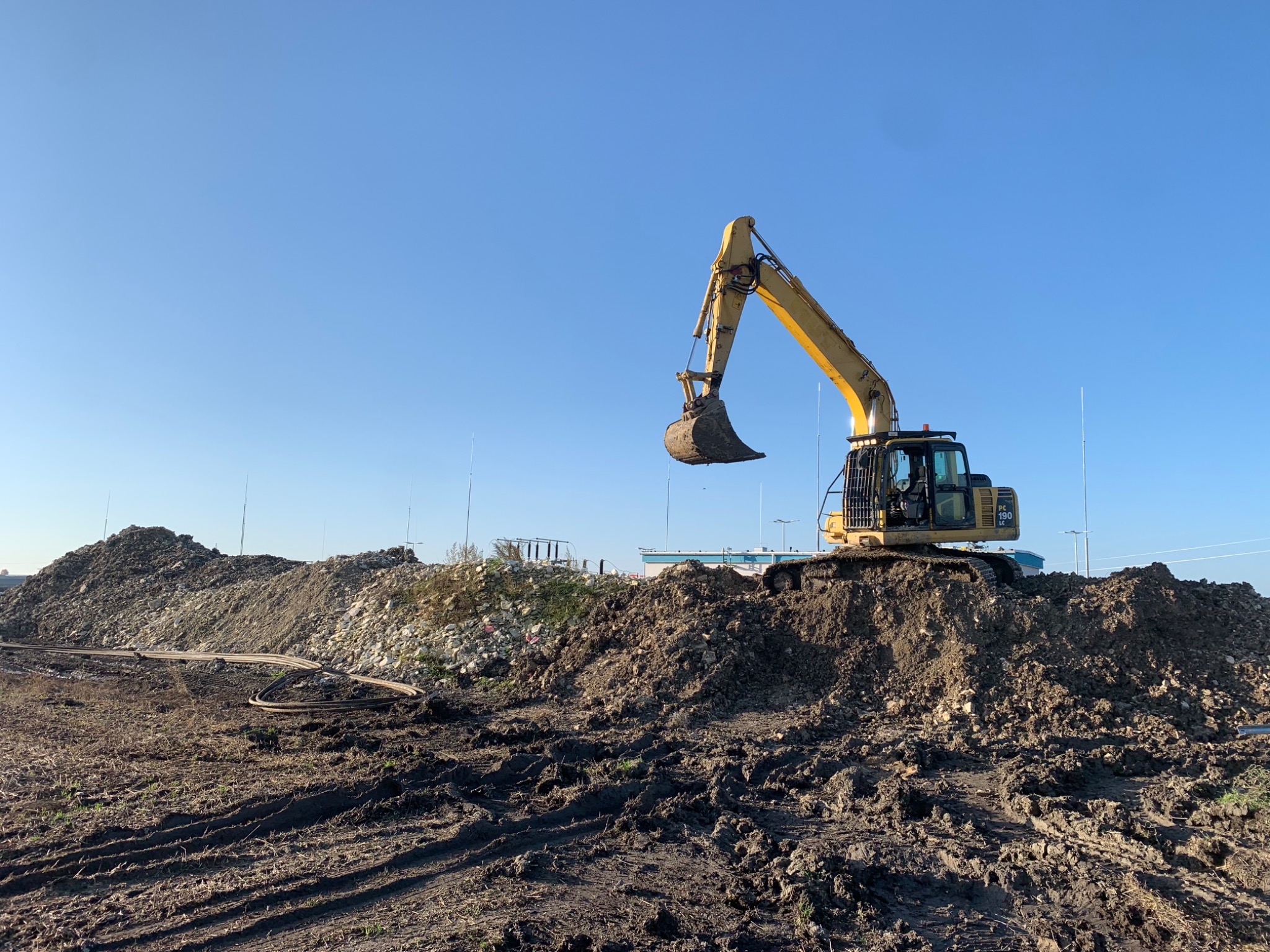 Żółta koparka na wzgórzu ziemi z widocznymi śladami opon i rurami w tle, błękitne niebo.