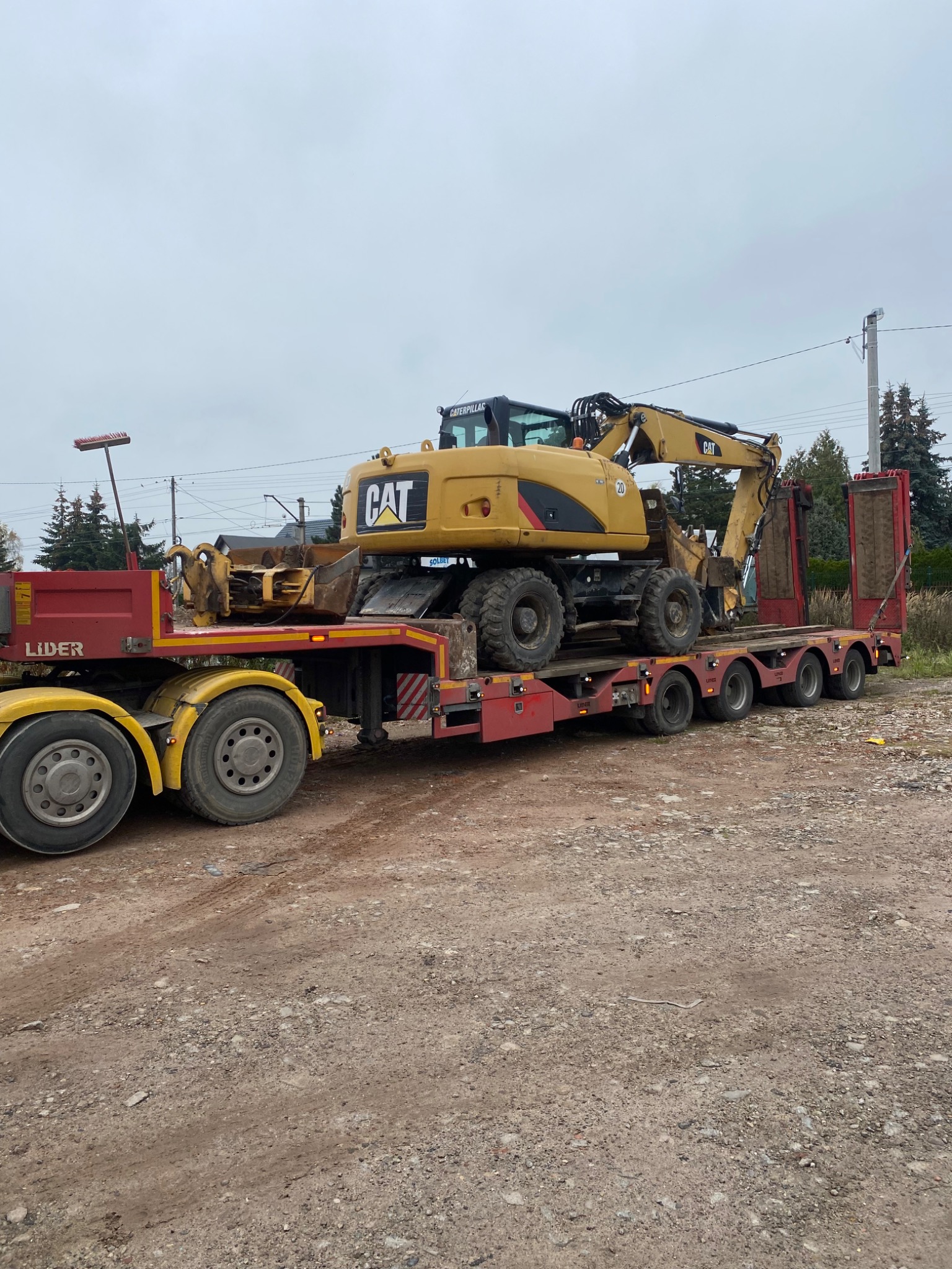 Żółta koparka kołowa marki CAT transportowana na niskopodwoziowej platformie z czerwoną kabiną ciężarówki na szutrowej drodze.
