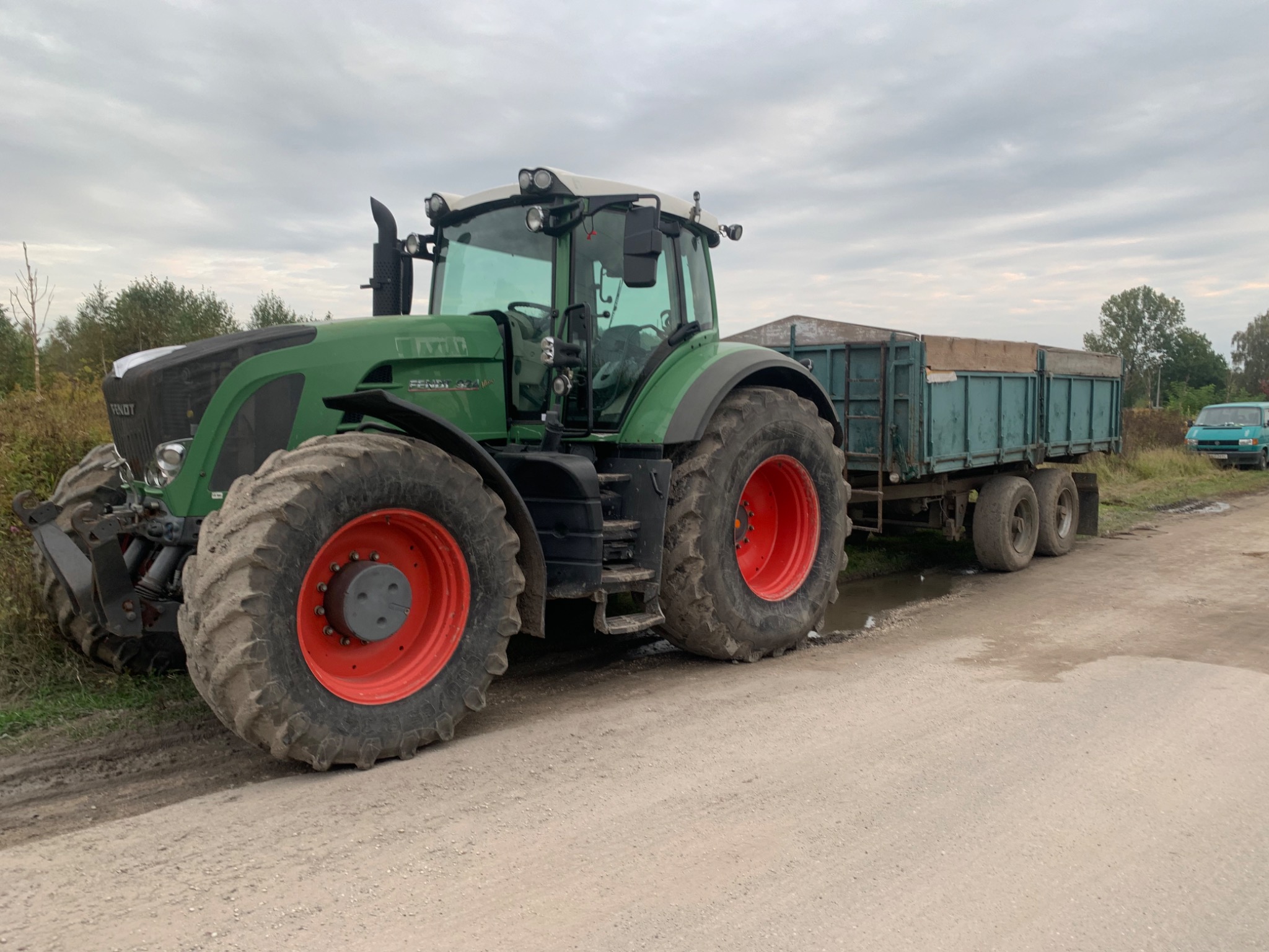 Zielony traktor Fendt z czerwoną felgą i przyczepą na polnej drodze, tło z drzewami i pochmurnym niebem.