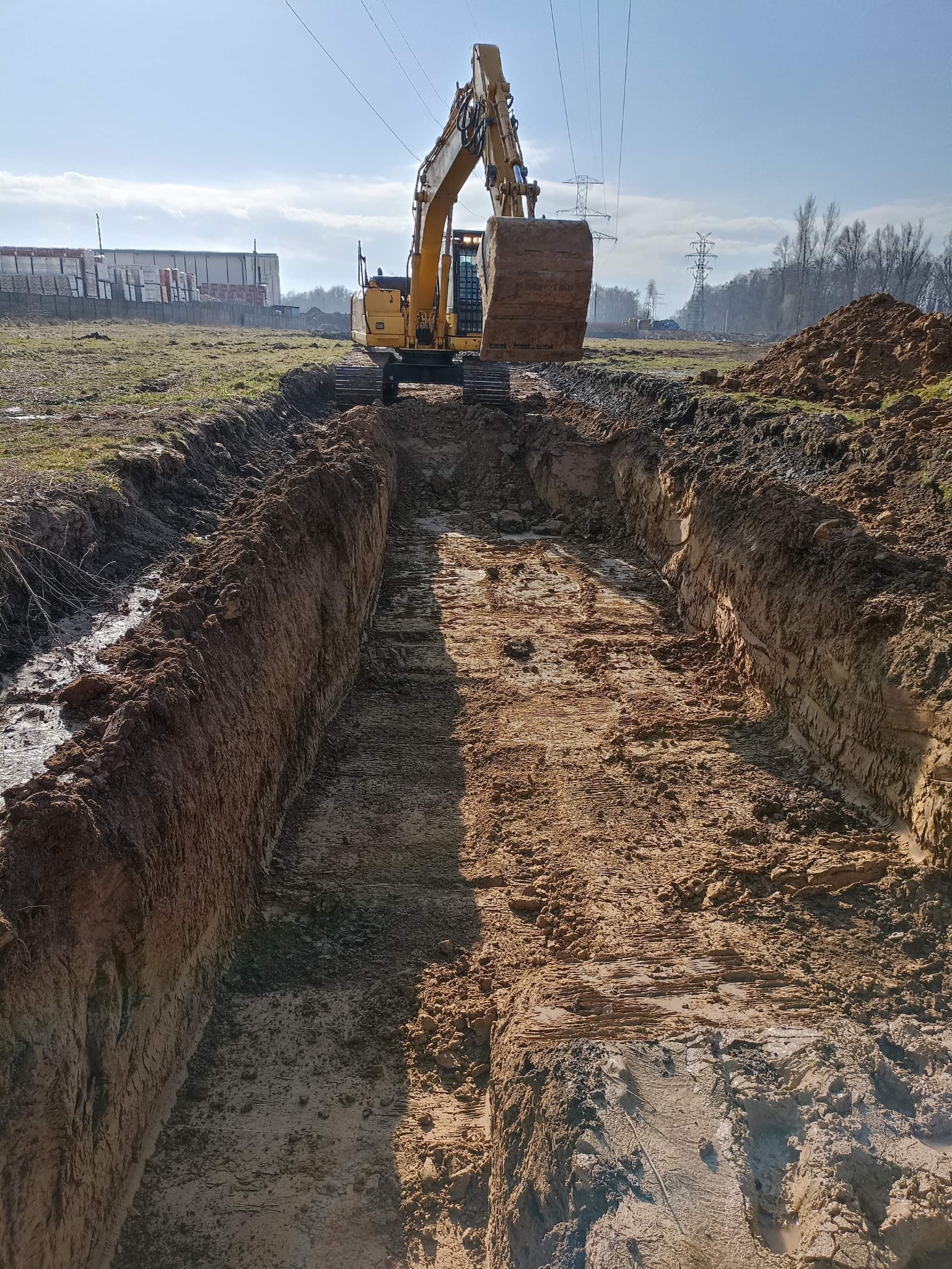 Żółta koparka na tle błękitnego nieba, wykonująca wykop liniowy w ziemi, widoczne ślady gąsienic i pryzma ziemi obok wykopu, w tle budynki przemysłowe i słupy wysokiego napięcia.