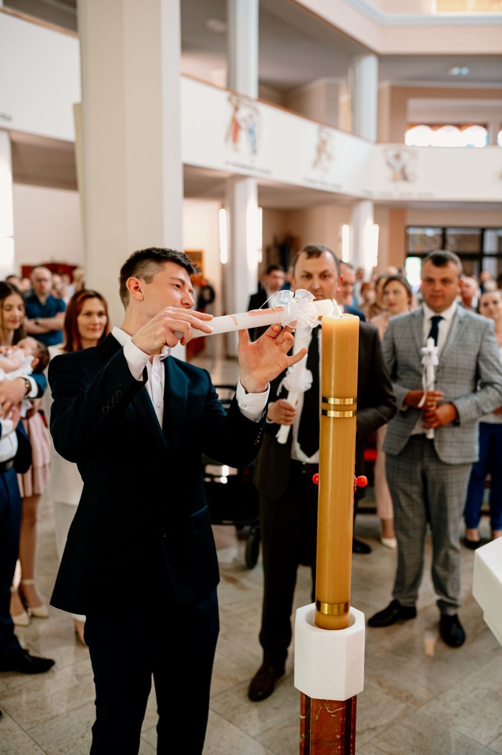 Mężczyzna w garniturze zapala świecę chrzcielną w kościele, w tle widoczni goście uroczystości.