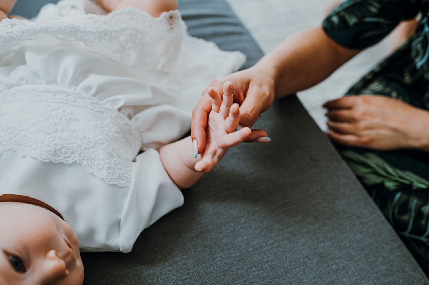 Dziecko w białej, koronkowej sukience leży na szarym tle, trzymając za rękę dorosłą osobę z niebieskim manicure.