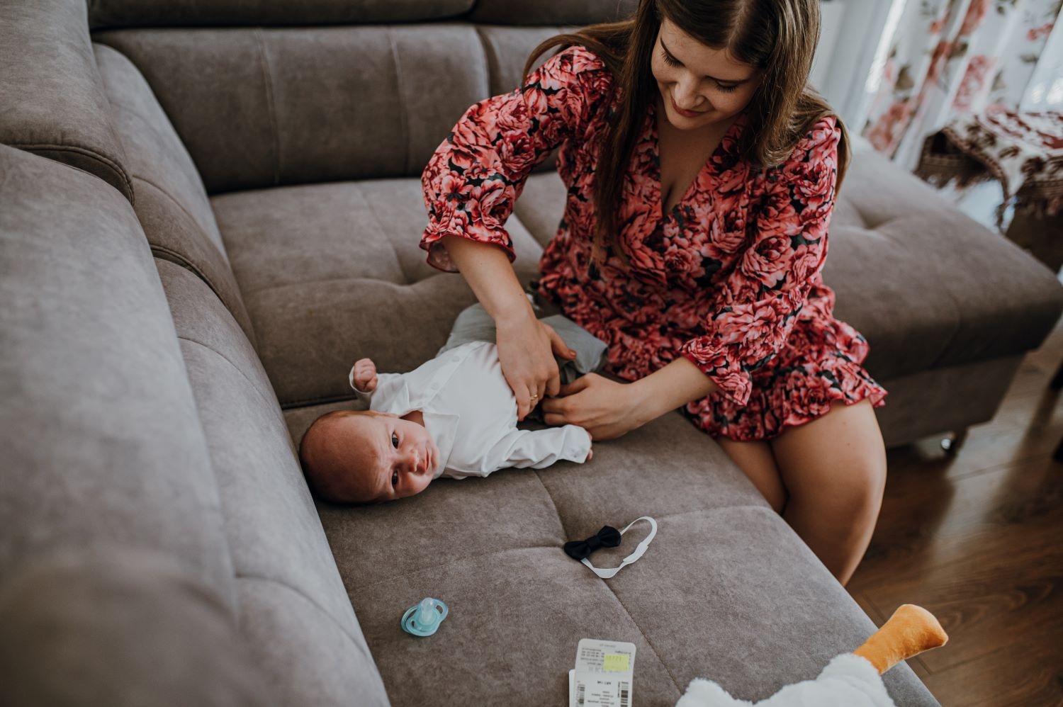 Mama w kwiecistej sukience ubiera leżącego na szarym fotelu niemowlaka, obok smoczek, muszka i pluszowa kaczka.