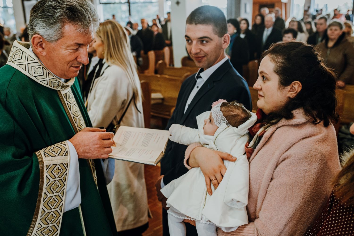 Ksiądz w zielonej szacie czyta z księgi podczas ceremonii, mama trzyma ubraną na biało dziewczynkę, tata patrzy z uśmiechem, w tle zgromadzeni ludzie w kościele.