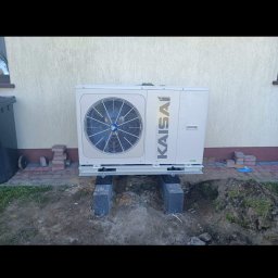 Nortmar Oddział Opole - Zewnętrzna jednostka pompy ciepła Kaisai zamontowana na betonowych blokach przed beżową ścianą budynku, widoczne kostki brukowe i ziemia.