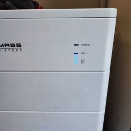 Nortmar Oddział Opole - Biały magazyn energii MASS STORE z diodami LED wskazującymi status i poziom naładowania (SOC), z fragmentem rękawicy roboczej w tle.