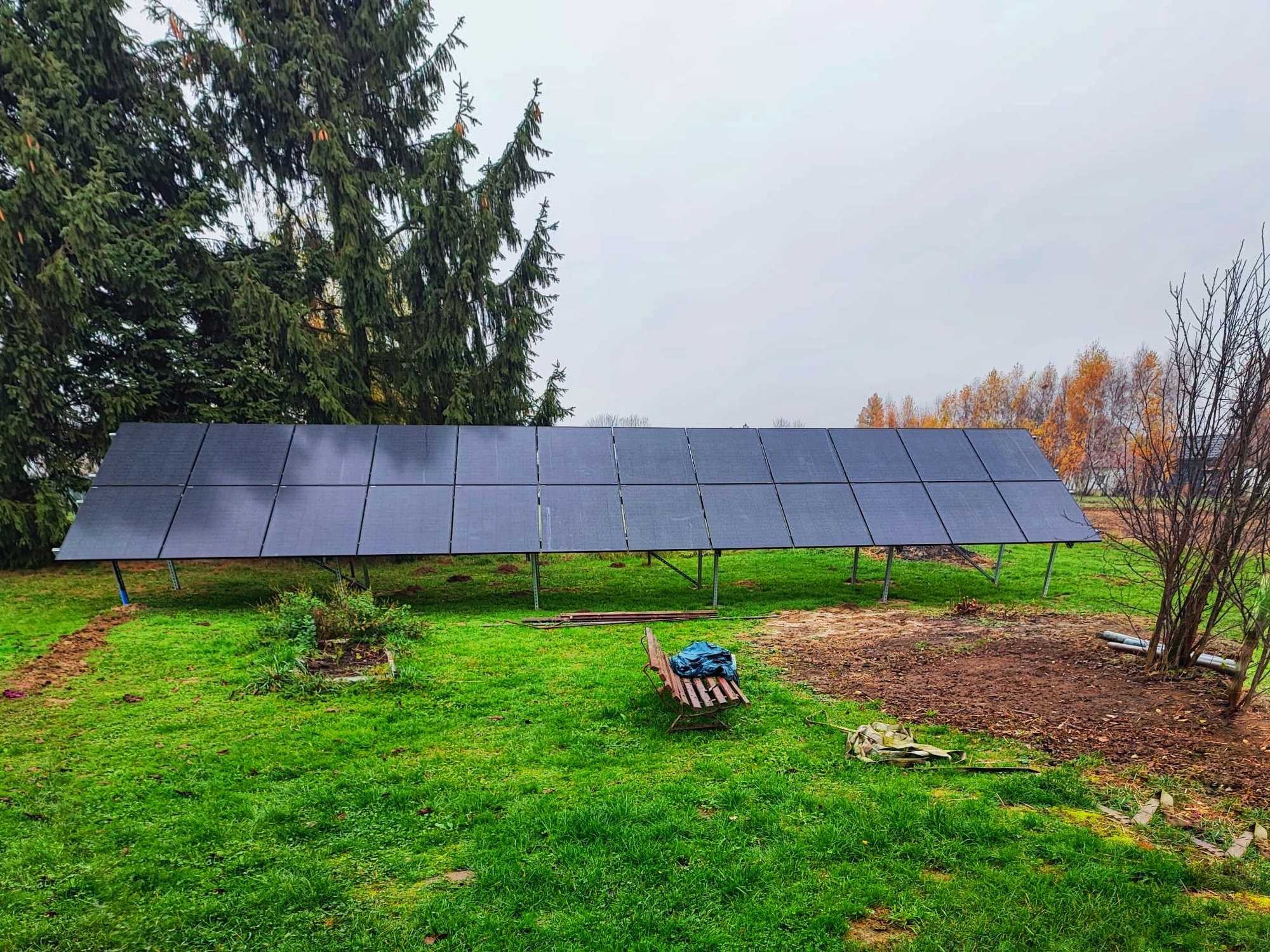 Rozległy panel słoneczny na metalowych wspornikach, zainstalowany na trawiastym terenie, z widocznym drewnianym siedziskiem i rozkopaną ziemią w tle drzew.