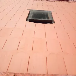 Czerwona dachówka ceramiczna, widok z góry na fragment dachu z oknem dachowym w czarnej ramie i barierką zabezpieczającą. Dachówka ułożona równo, widać spoiny.