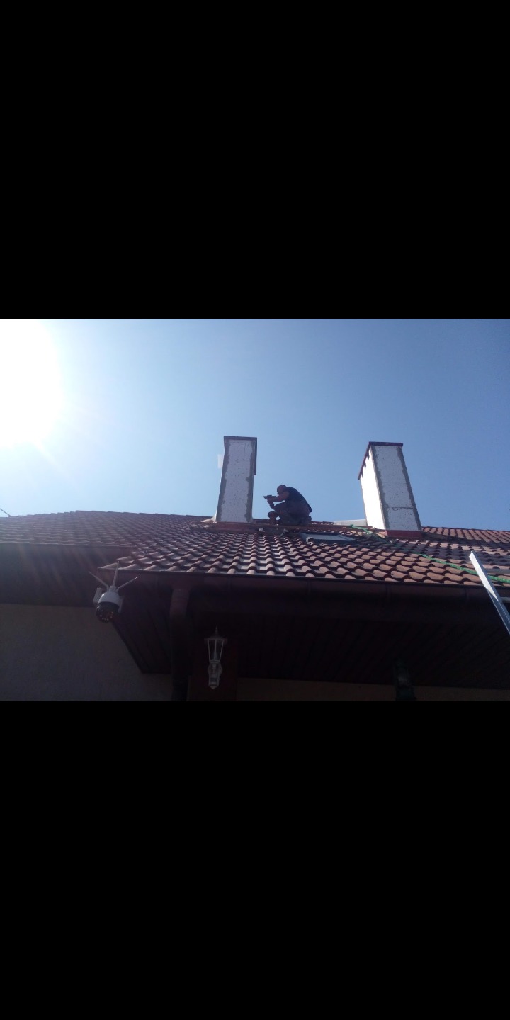 Pracownik na dachu z dachówki ceramicznej, montujący obudowę komina z płyt izolacyjnych, widok z dołu na tle błękitnego nieba.