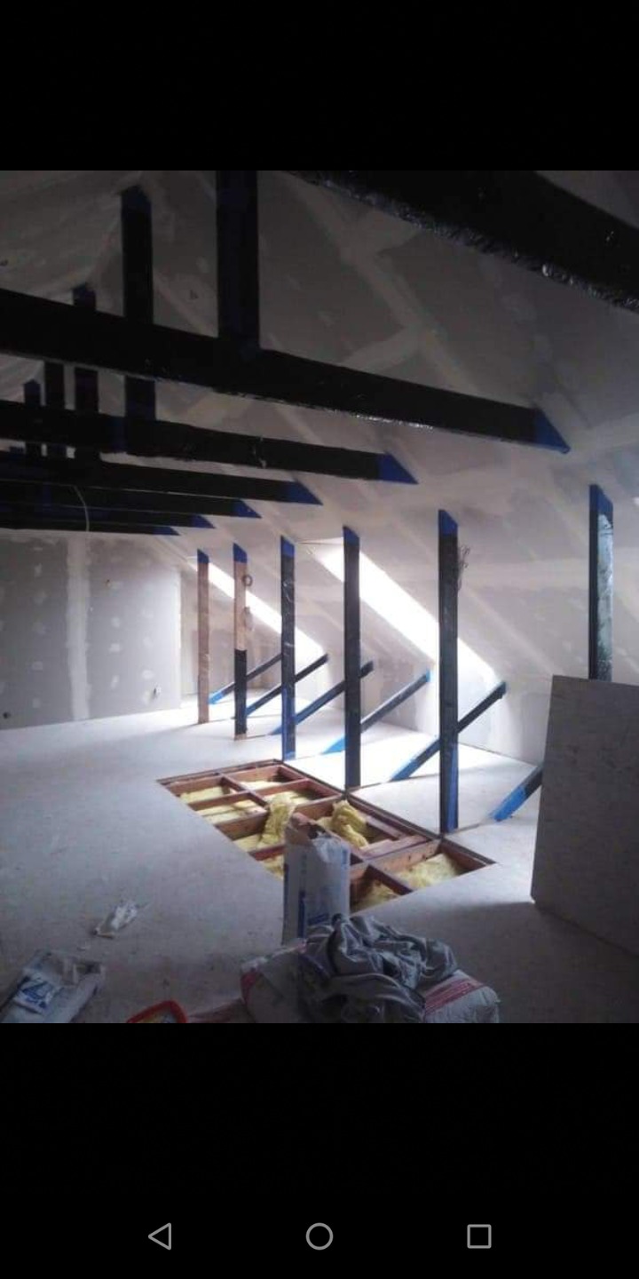 Poddasze w trakcie adaptacji: widoczne ocieplenie w podłodze, konstrukcja wsporcza z belek, częściowo wykończone ściany płytami gipsowo-kartonowymi, worki z materiałami budowlanymi.