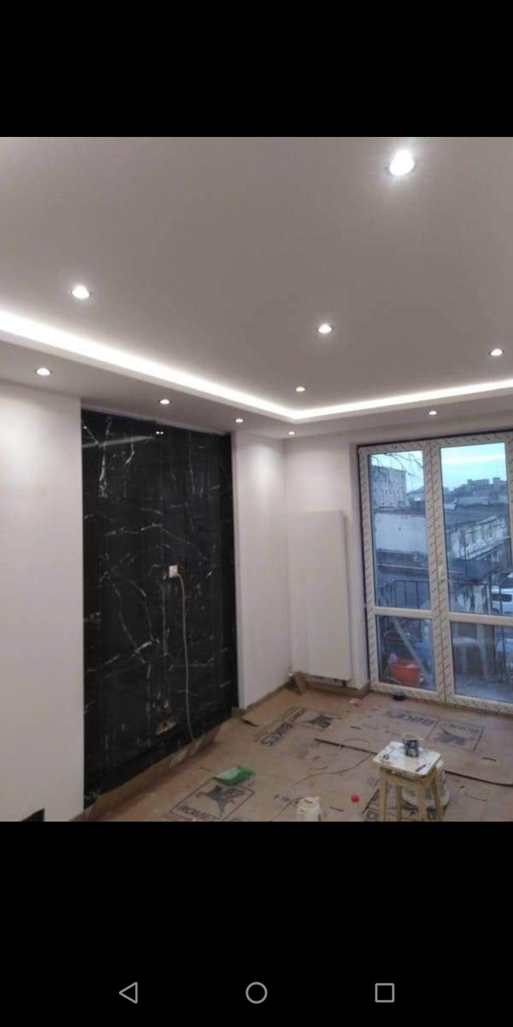 Wnętrze pokoju w trakcie remontu: czarna ściana z marmurowym wzorem, podwieszany sufit z oświetleniem LED, zabezpieczona podłoga kartonem, okno z widokiem na miasto.