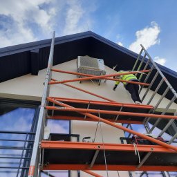 KlimaHome Sp. z o.o. - Pracownik w żółtej kamizelce na rusztowaniu montuje jednostkę zewnętrzną klimatyzacji na ścianie domu z ciemnym dachem i dużym oknem.