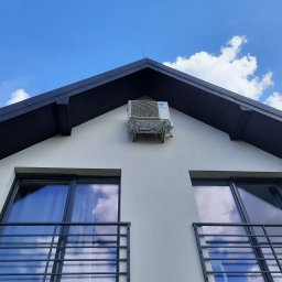 KlimaHome Sp. z o.o. - Biały klimatyzator zamontowany na ścianie szczytowej domu z czarnym dachem na tle błękitnego nieba z chmurami, widok z dołu na balkon z metalową balustradą.