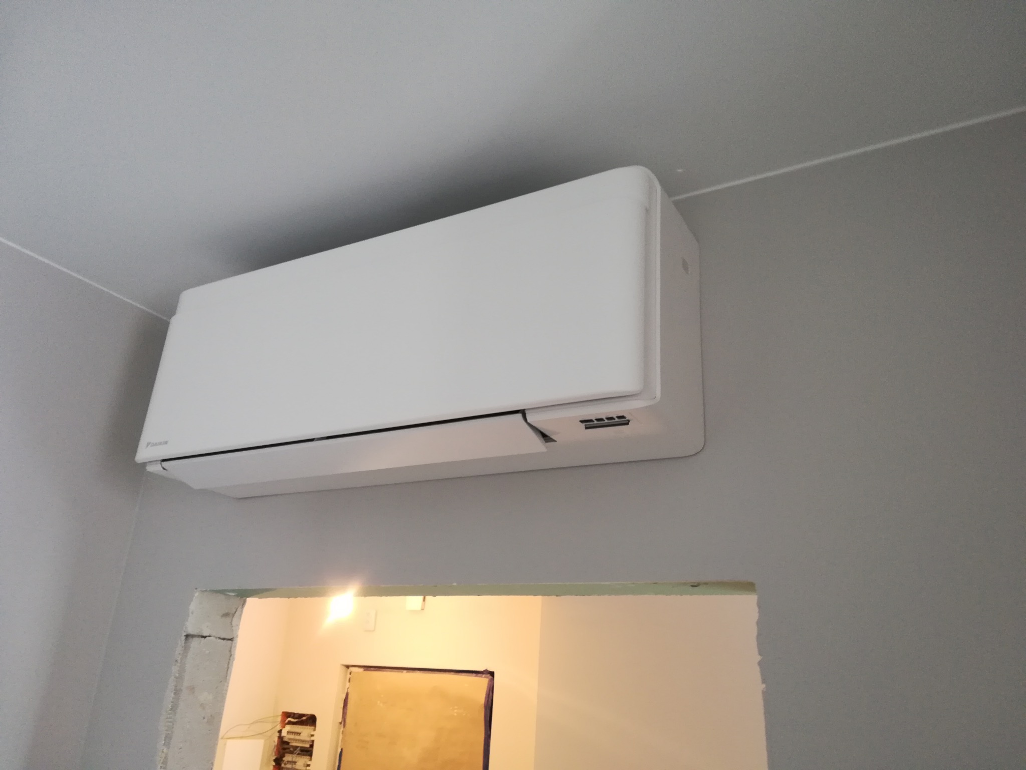 Biała jednostka klimatyzacji Daikin zamontowana na szarej ścianie nad otworem drzwiowym w trakcie remontu.