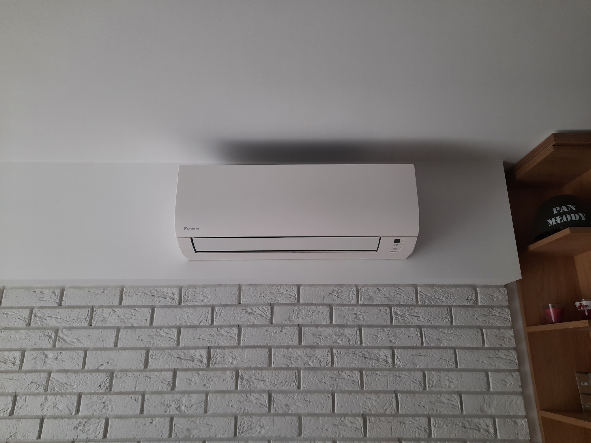 Biały klimatyzator Daikin zamontowany na białej ścianie z cegły dekoracyjnej, widoczny fragment drewnianego regału z dekoracjami i czapką z napisem.