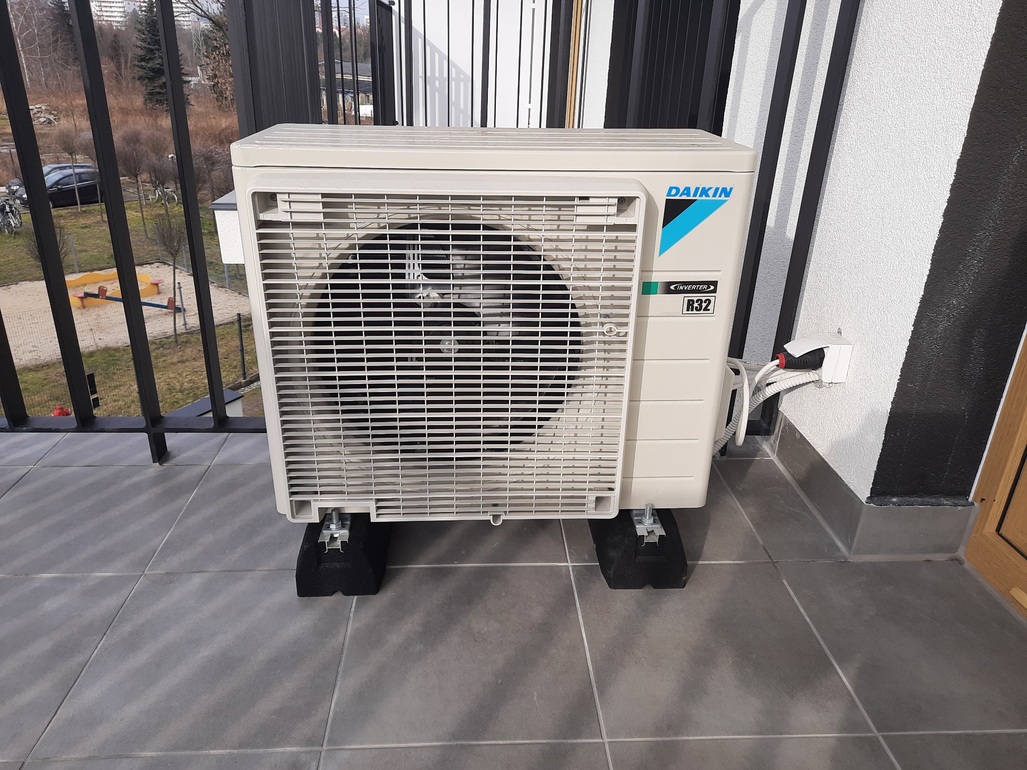 Jednostka zewnętrzna klimatyzatora Daikin zamontowana na balkonie z widokiem na plac zabaw i drzewa.