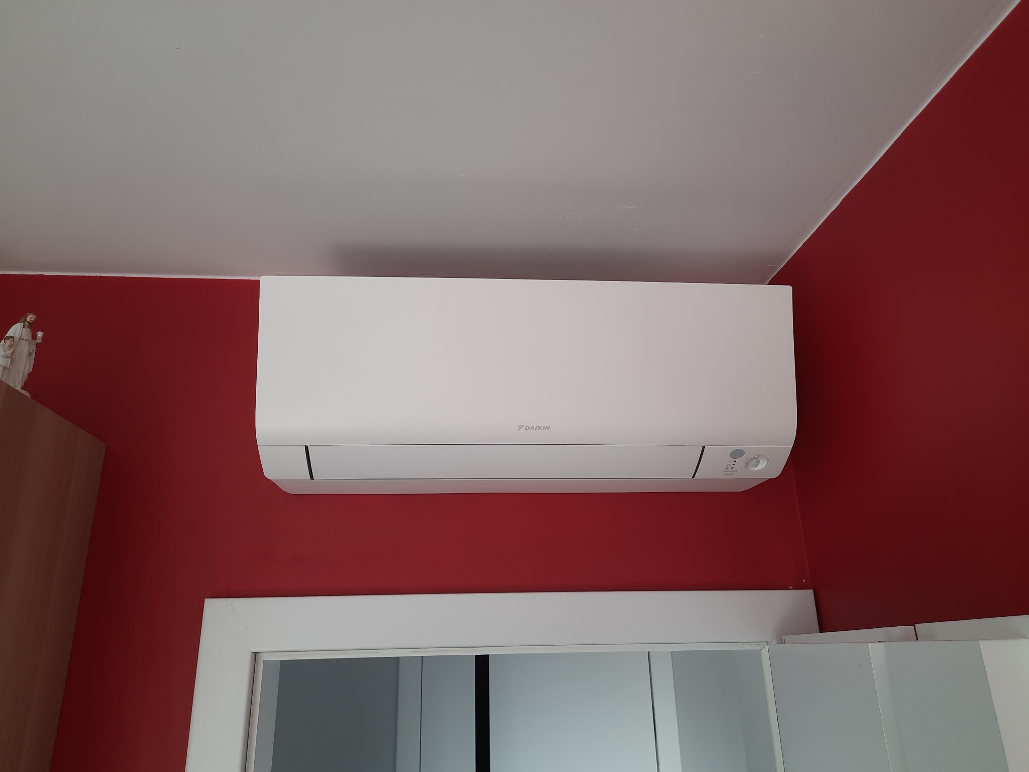 Biała klimatyzacja Daikin zamontowana na ścianie pomalowanej na intensywny czerwony kolor, widoczna nad białą ramą drzwi i fragmentem szafki z figurką.
