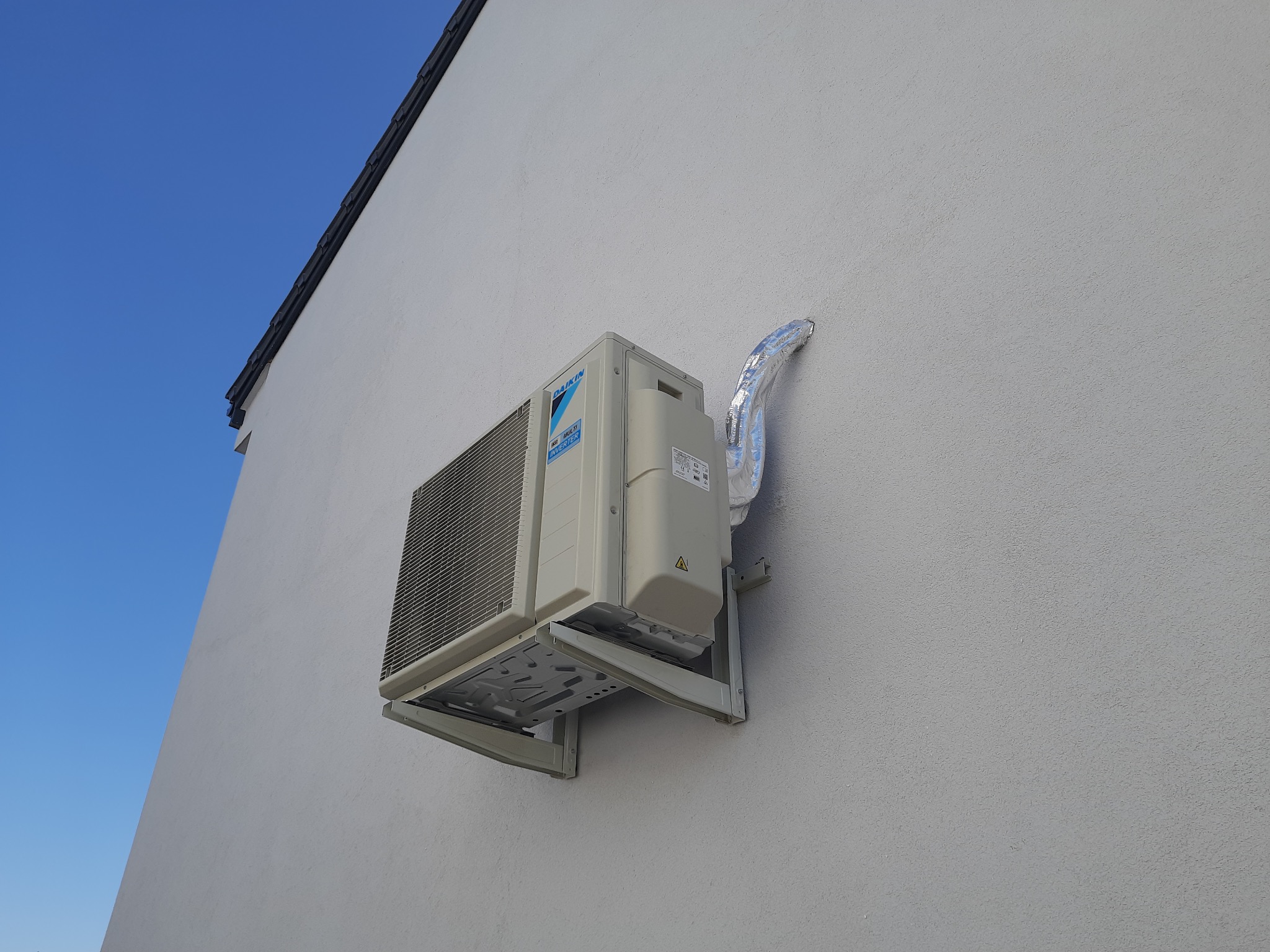 Zewnętrzna jednostka klimatyzacji Daikin zamontowana na jasnej, teksturowanej ścianie budynku z widocznym fragmentem dachu i błękitnym niebem.