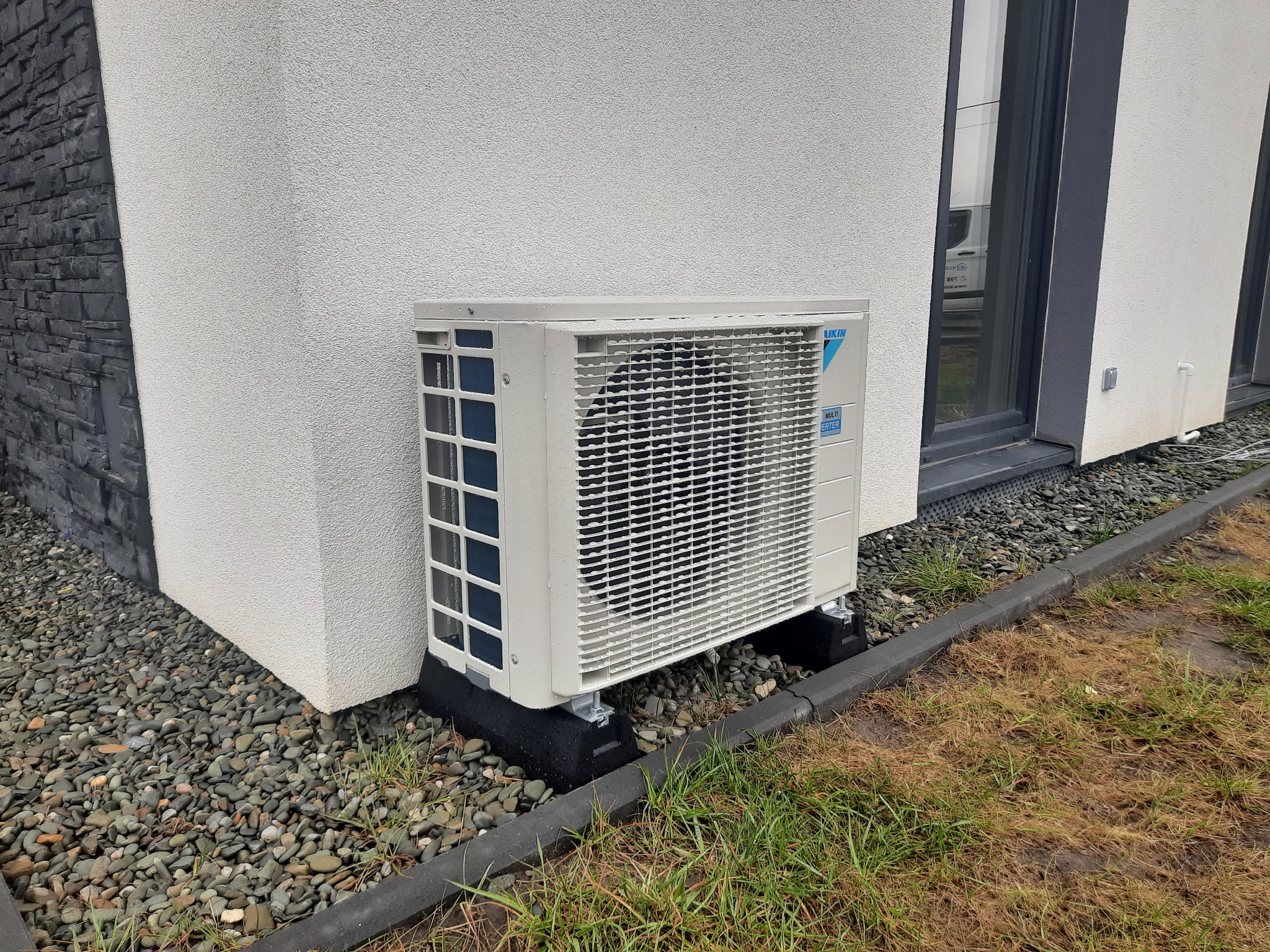 Zewnętrzna jednostka klimatyzacji Daikin Multi Inverter zamontowana przy ścianie domu z elewacją w kolorze białym i elementami z szarej cegły, posadowiona na czarnych, gumowych podkładkach...