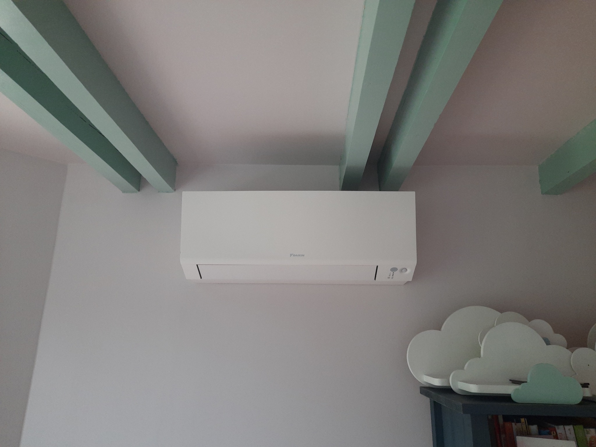 Biały klimatyzator Daikin zamontowany na szarej ścianie w pokoju dziecięcym, widoczne drewniane, pomalowane na zielono belki stropowe i dekoracja w kształcie chmur na szafce.