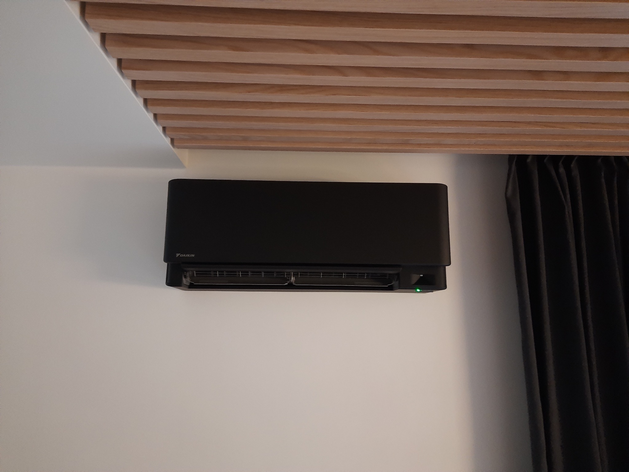 Czarna jednostka klimatyzacji Daikin zamontowana na białej ścianie w pomieszczeniu z drewnianym sufitem lamelowym i zasłoną w tle.