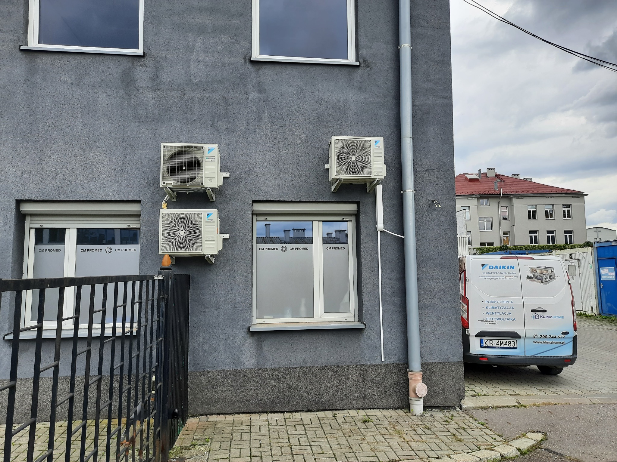 Szara ściana budynku w Krakowie z czterema zamontowanymi klimatyzatorami marki Daikin, widoczne okna z roletami i logo firmy CM Promed, a w tle zaparkowany bus firmy KlimaHome z reklamą usług.