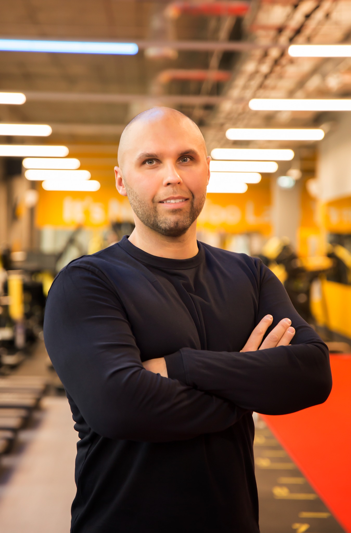 Portret uśmiechniętego mężczyzny w czarnej koszulce z długim rękawem, stojącego na tle siłowni z czerwonym pasem na podłodze i żółtymi elementami wyposażenia.