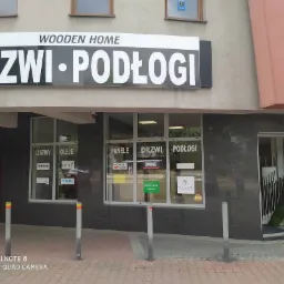 Fasada sklepu Wooden Home z dużym szyldem DRZWI PODŁOGI, okna z naklejkami reklamowymi listew, paneli, drzwi i podłóg, oraz otwarte drzwi wejściowe.