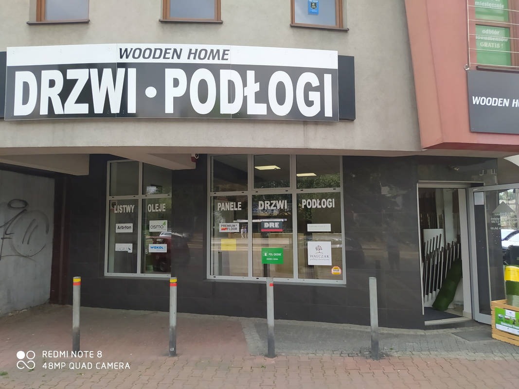 Fasada sklepu Wooden Home z dużym szyldem DRZWI PODŁOGI, okna z naklejkami reklamowymi listew, paneli, drzwi i podłóg, oraz otwarte drzwi wejściowe.