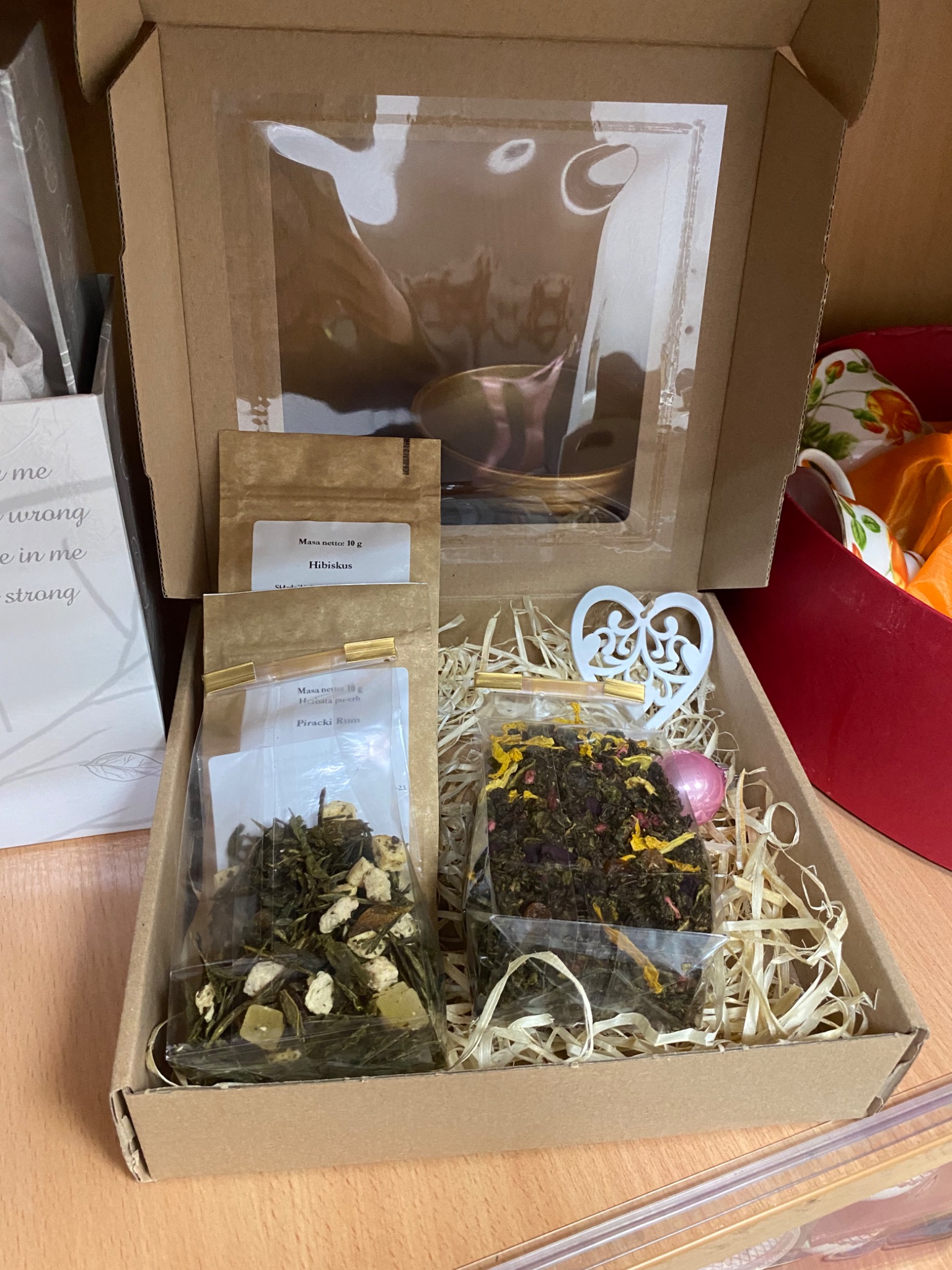 Otwarty kartonowy box wypełniony siankiem, zawierający herbaty liściaste w przezroczystych torebkach z etykietami, ozdobę w kształcie serca i małą różową bombkę. W tle widoczna filiżanka.