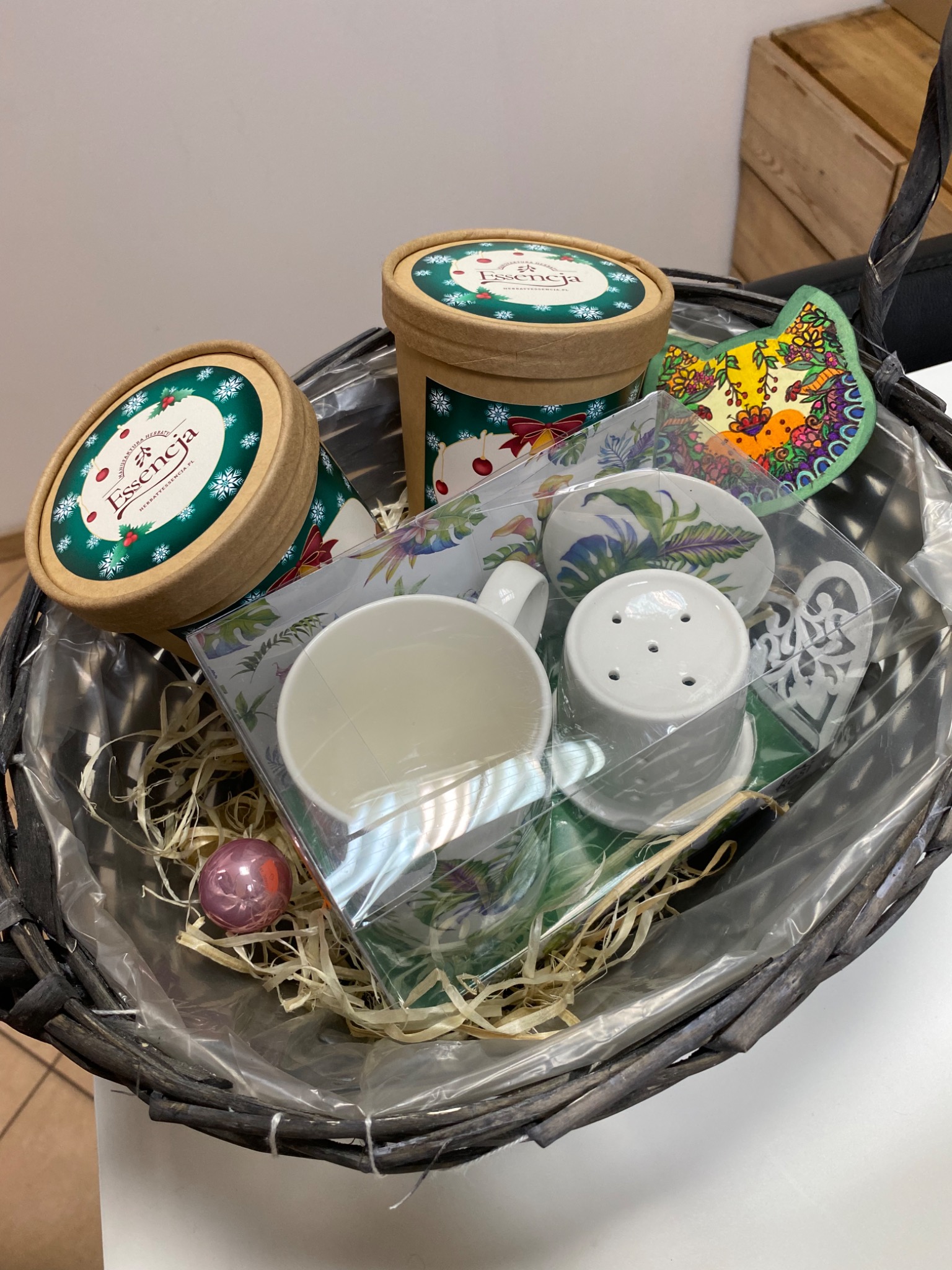 Ręcznie robiony kosz upominkowy z herbatami Essencja, ceramiczną filiżanką i ozdobami, zapakowany w celofan i wypełniony siankiem, oferowany przez firmę ze Szczecina.