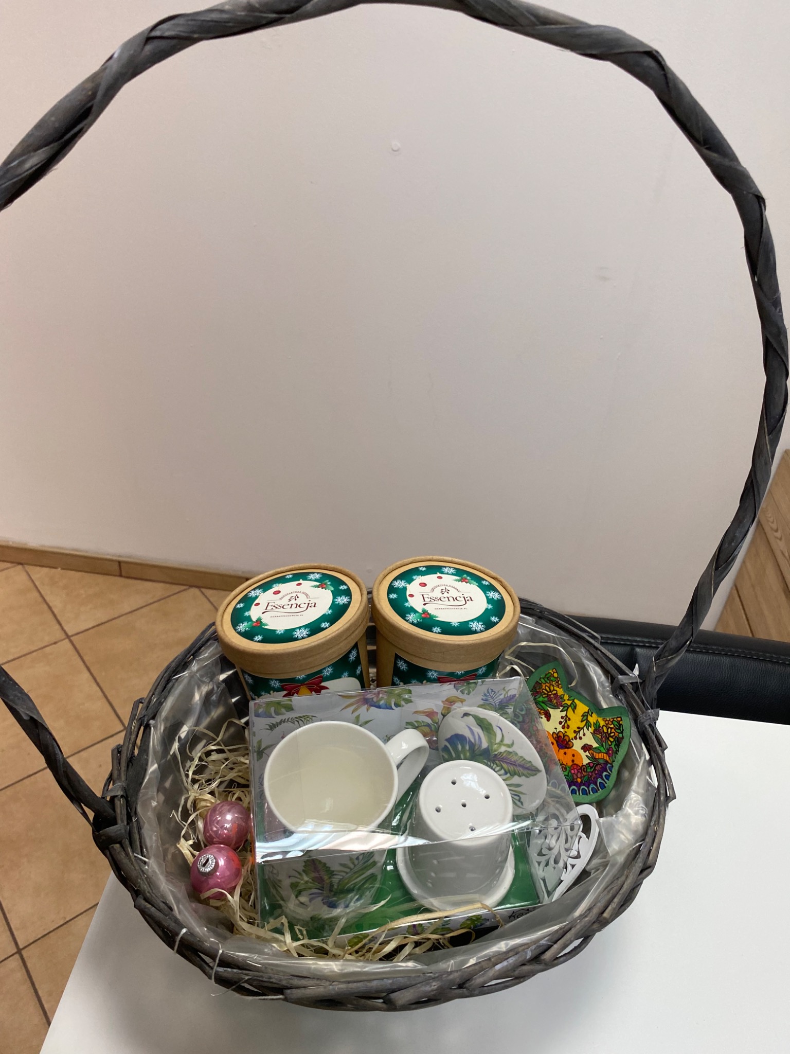 Szary kosz prezentowy z dwoma okrągłymi pojemnikami z zielonymi wieczkami z napisem Essencja, filiżanką, spodkiem, cukierniczką, podkładką z motywem ludowym i dwoma różowymi bombkami, wypełniony...