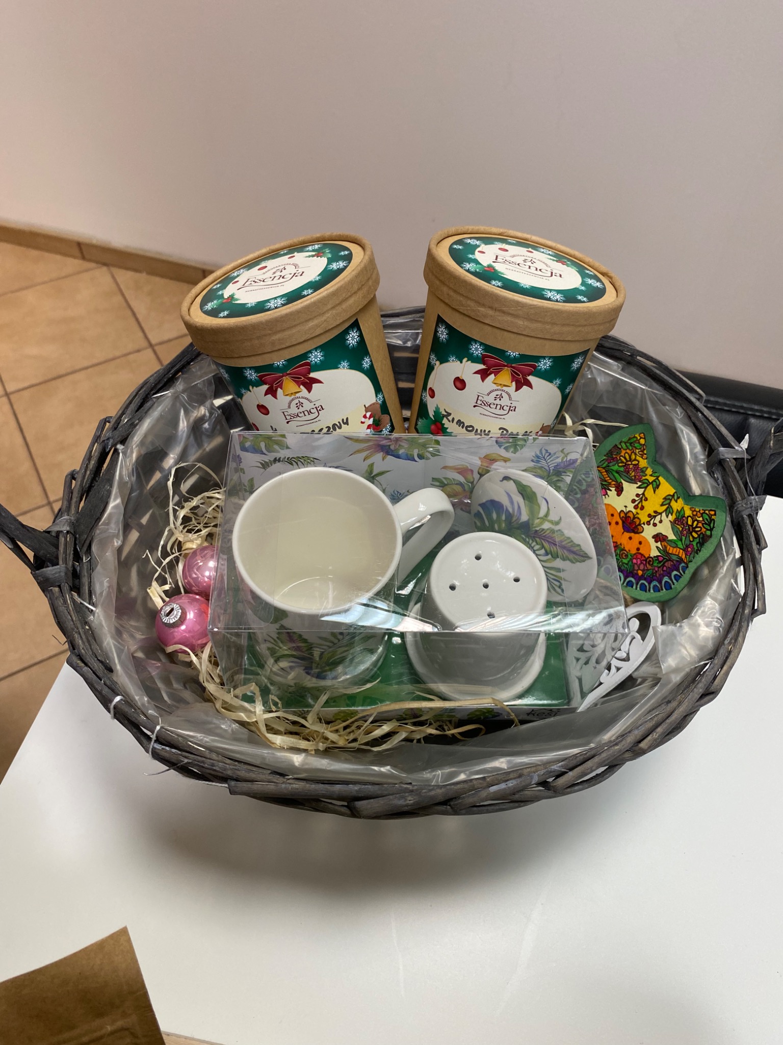 Szary kosz wiklinowy z dwoma okrągłymi pudełkami z motywem świątecznym, zestawem do herbaty z porcelany w przezroczystym pudełku, ozdobami choinkowymi i drewnianą ozdobą w kształcie liścia.