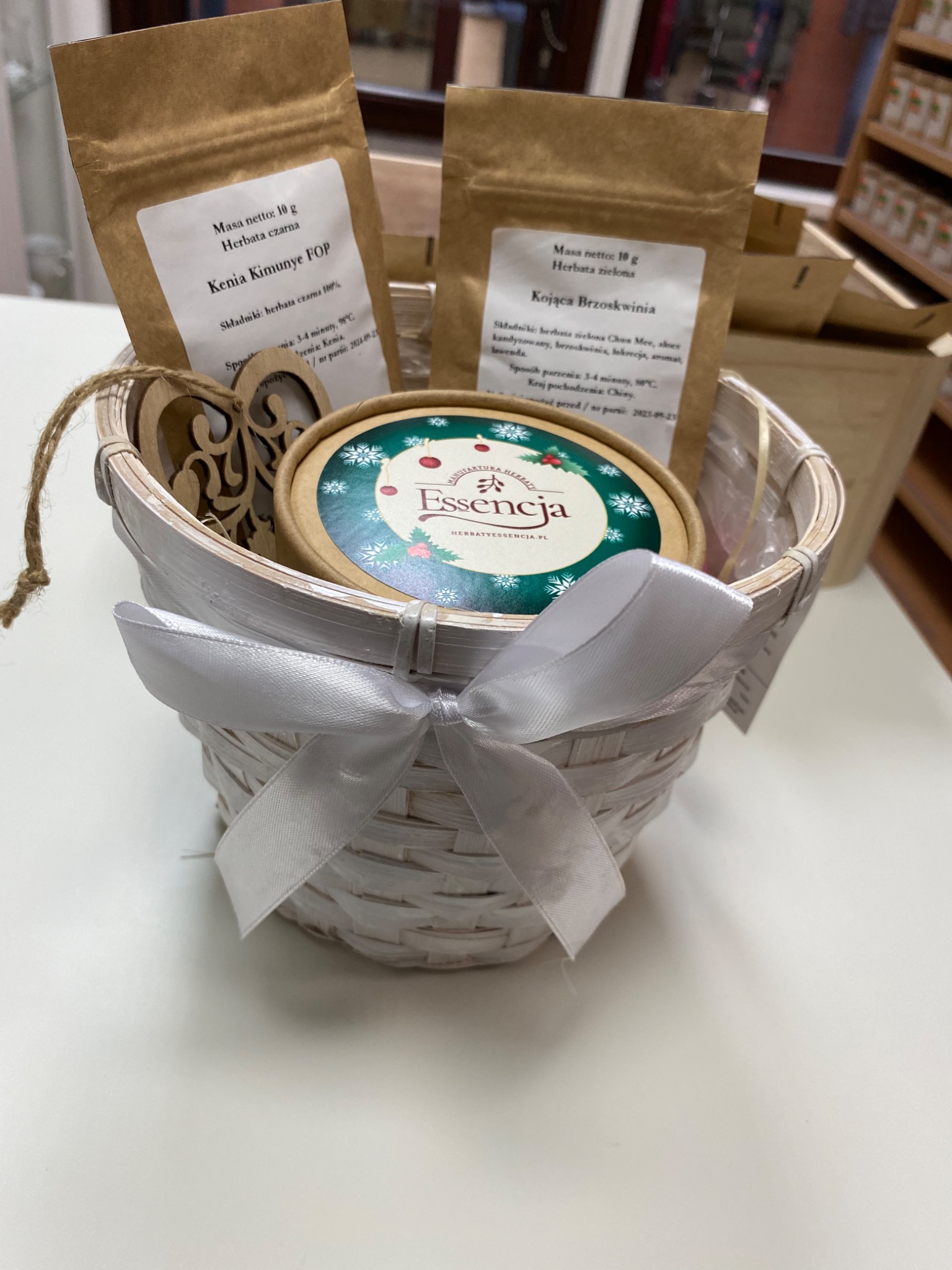 Biały, pleciony koszyk z jasną wstążką, zawierający dwa opakowania herbaty (czarnej i zielonej) oraz okrągłe pudełko z logo 'Essencja'. W koszyku widoczna drewniana ozdoba w kształcie serca. Całość...