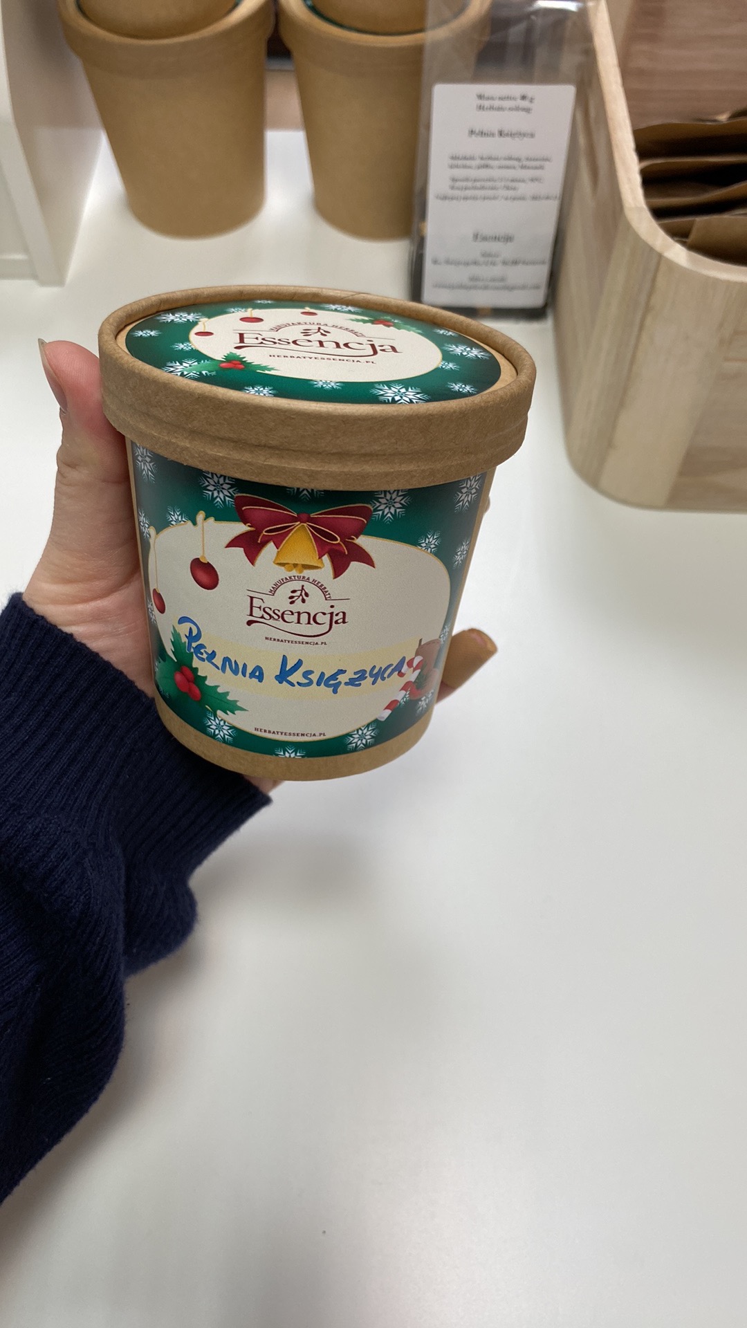 Ręka trzyma zielony pojemnik z brązową pokrywką, świątecznym motywem i napisem 'Essencja Pełnia Księżyca', w tle widoczne inne pojemniki i drewniany koszyk.