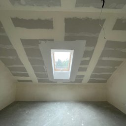 GrafRemo - Poddasze z oknem dachowym w trakcie prac wykończeniowych: widoczne łączenia płyt na suficie i ścianach, częściowo pokryte masą szpachlową, wystający kabel elektryczny, surowa betonowa podłoga.