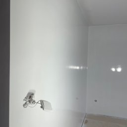 GrafRemo - Białe ściany i sufit w rogu pokoju po świeżym malowaniu. Widoczne gniazdko elektryczne i fragment podłogi. Nowoczesne wykończenie wnętrza, minimalistyczny styl.