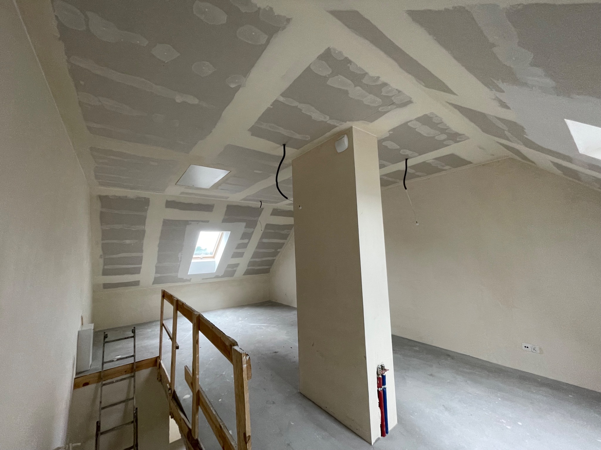 Poddasze w trakcie prac wykończeniowych: widoczne płyty na suficie, wstępnie zagruntowane ściany, pion instalacyjny z rurami, drabina i okno dachowe.