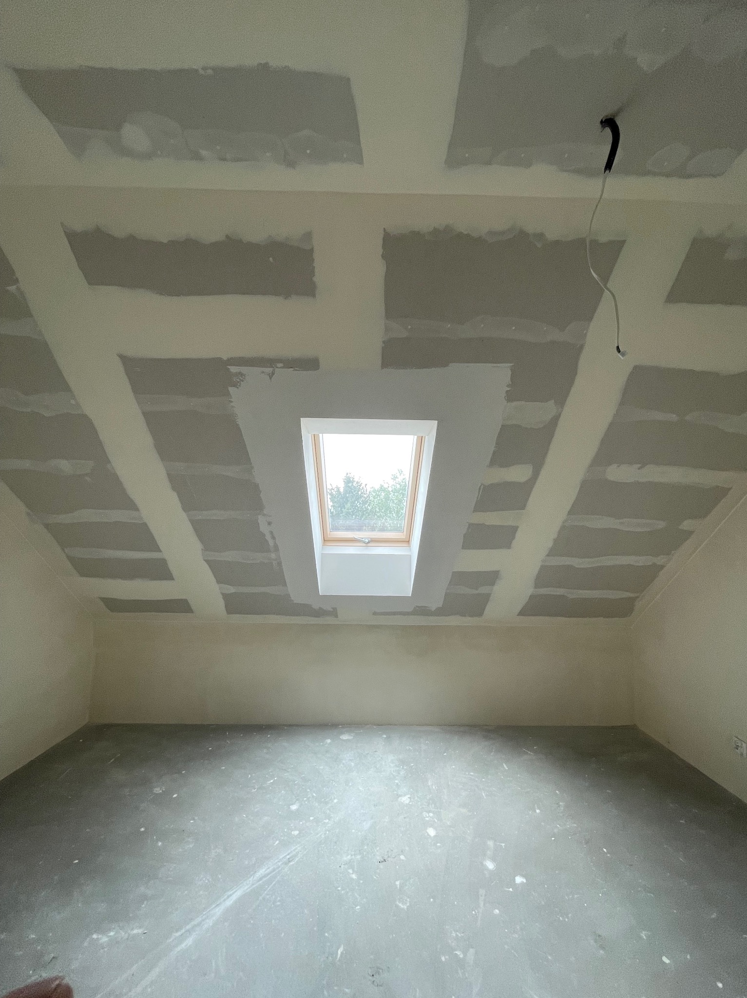 Poddasze z oknem dachowym w trakcie prac wykończeniowych: widoczne łączenia płyt na suficie i ścianach, częściowo pokryte masą szpachlową, wystający kabel elektryczny, surowa betonowa podłoga.