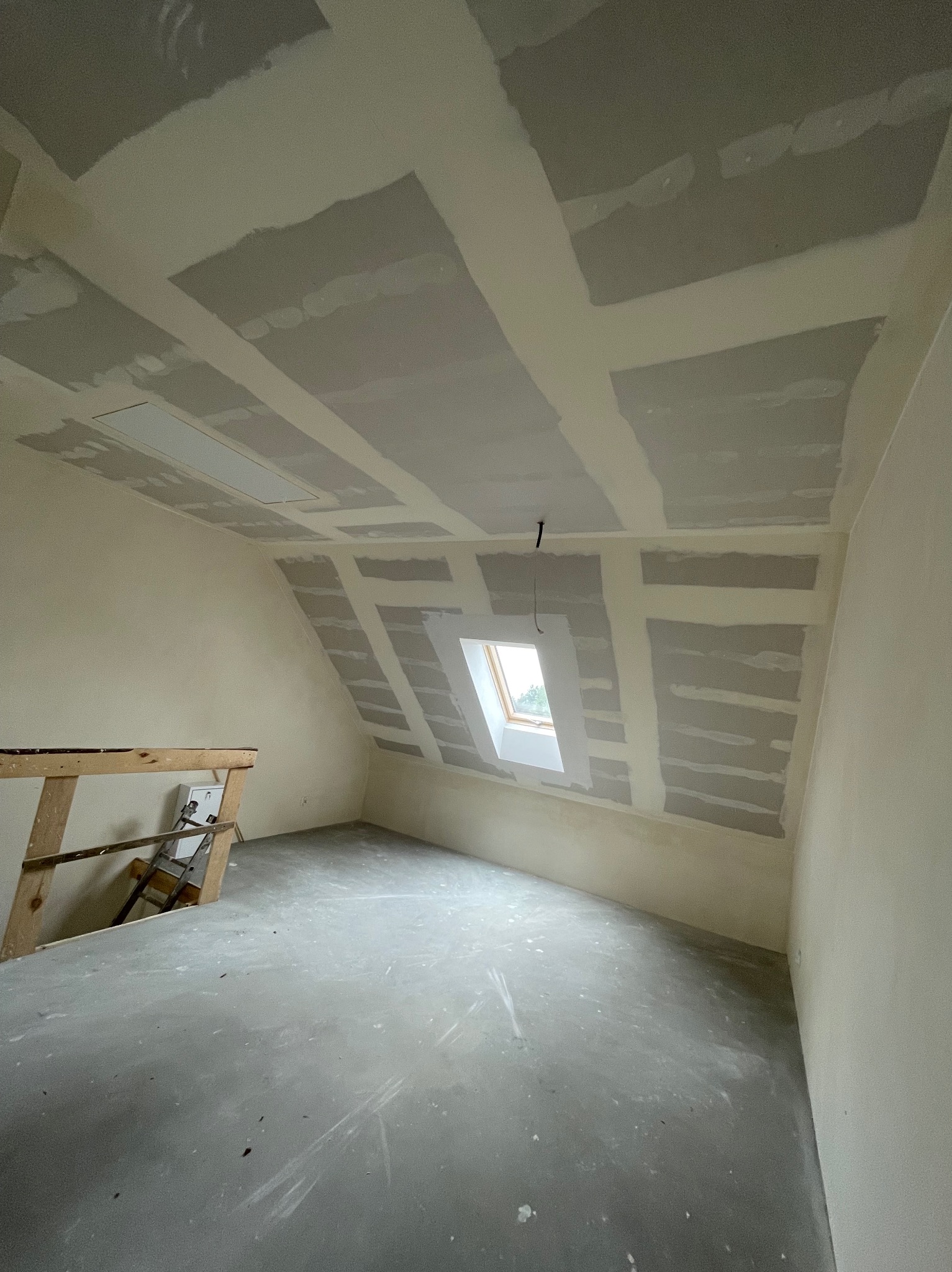 Poddasze w trakcie prac wykończeniowych: zamontowane płyty na skosach dachu i suficie z widocznymi łączeniami, okno dachowe w skosie, surowa podłoga, widoczny fragment drewnianego rusztowania.
