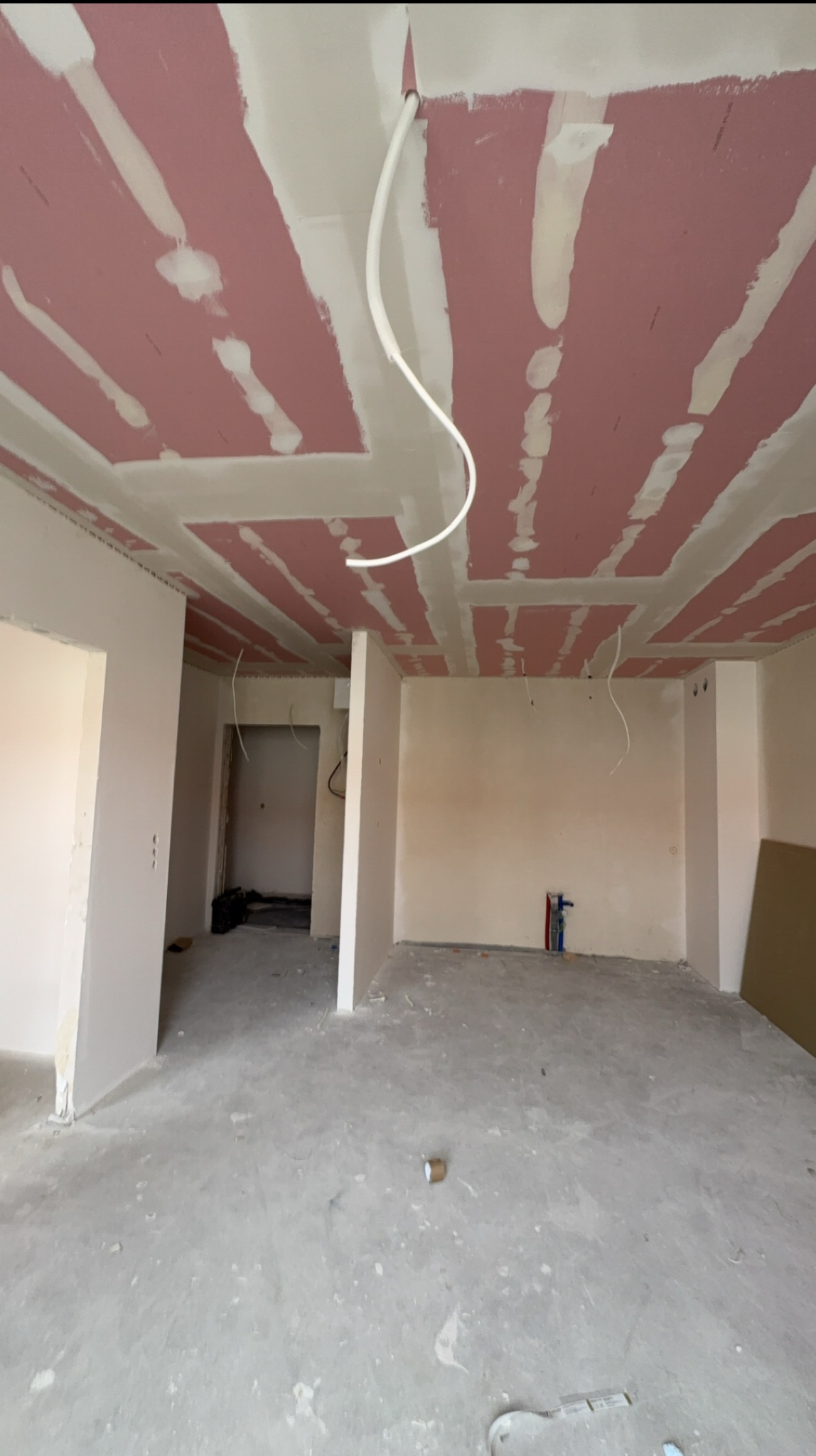 Wnętrze w trakcie prac: sufit z płyt gipsowych, widoczne łączenia i instalacje. Ściany przygotowane do dalszych prac wykończeniowych. Surowy, betonowy wygląd podłogi.