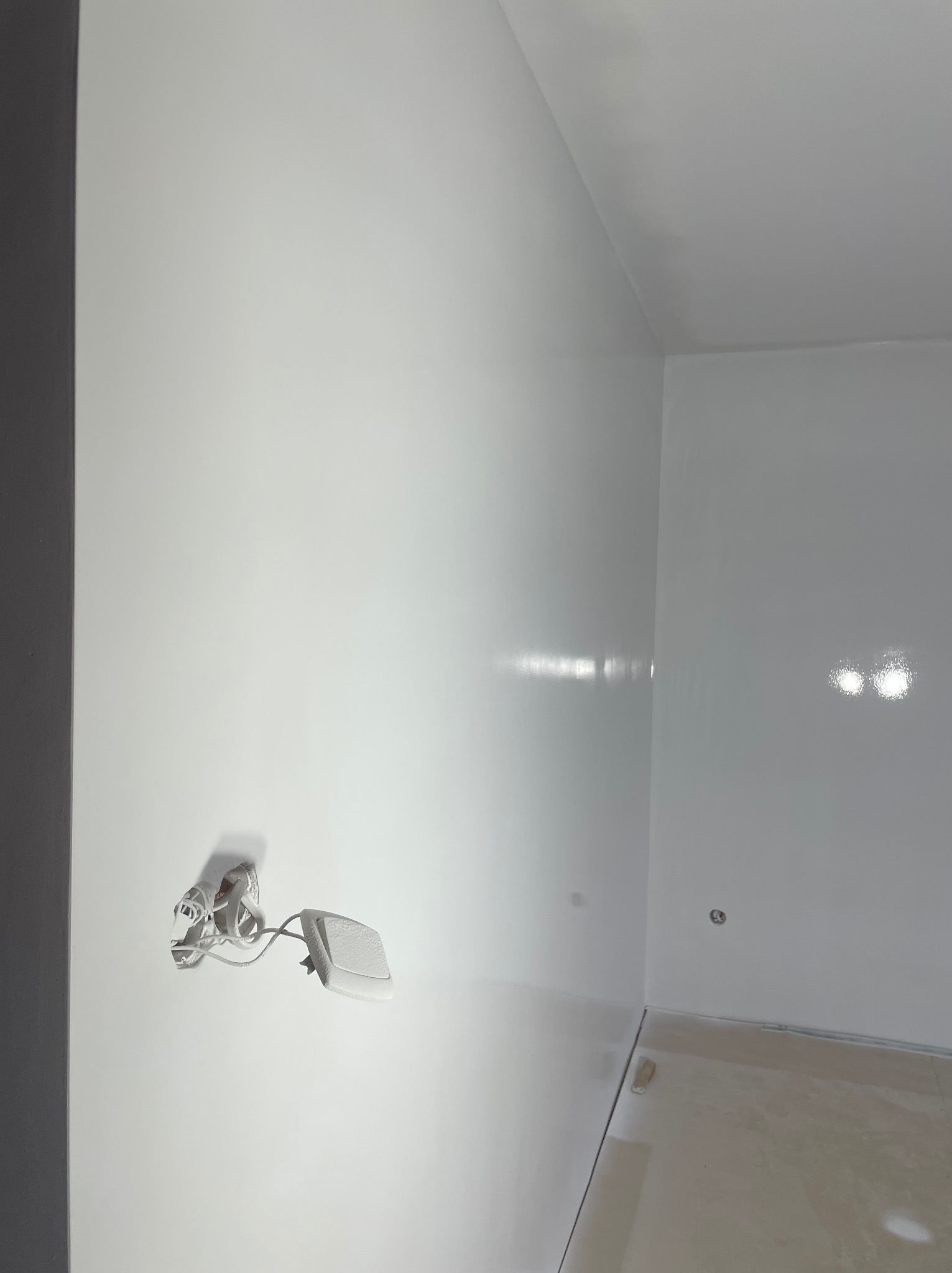 Białe ściany i sufit w rogu pokoju po świeżym malowaniu. Widoczne gniazdko elektryczne i fragment podłogi. Nowoczesne wykończenie wnętrza, minimalistyczny styl.