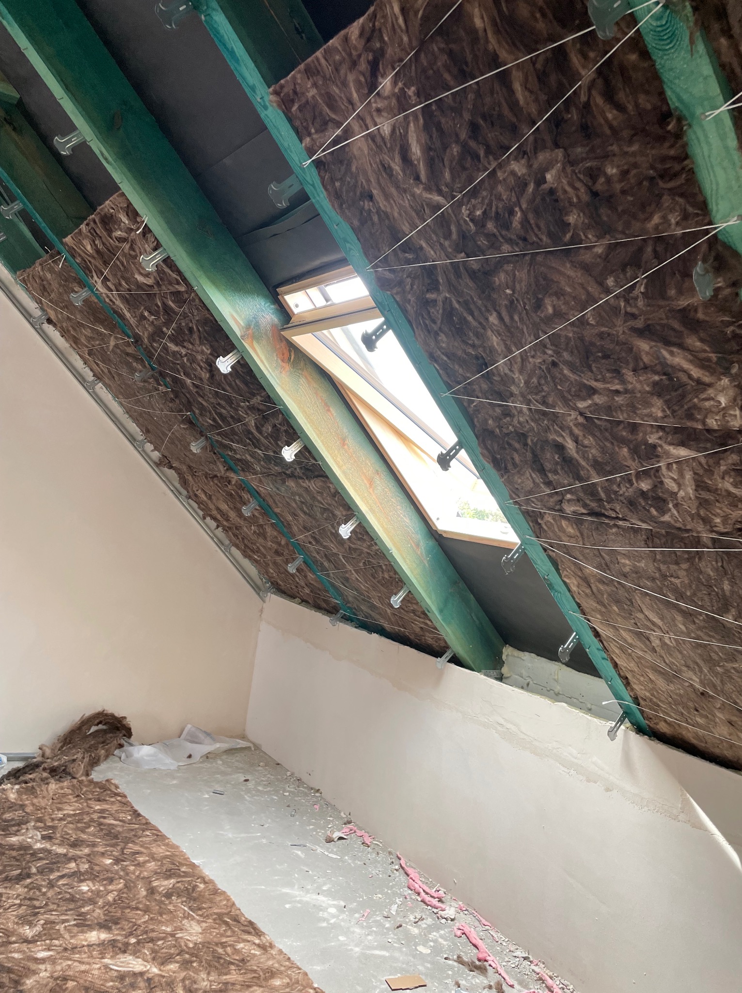 Poddasze z oknem dachowym w trakcie prac. Widoczne ocieplenie wełną mineralną, zielone belki konstrukcyjne i białe ściany. Częściowo wykończona powierzchnia.